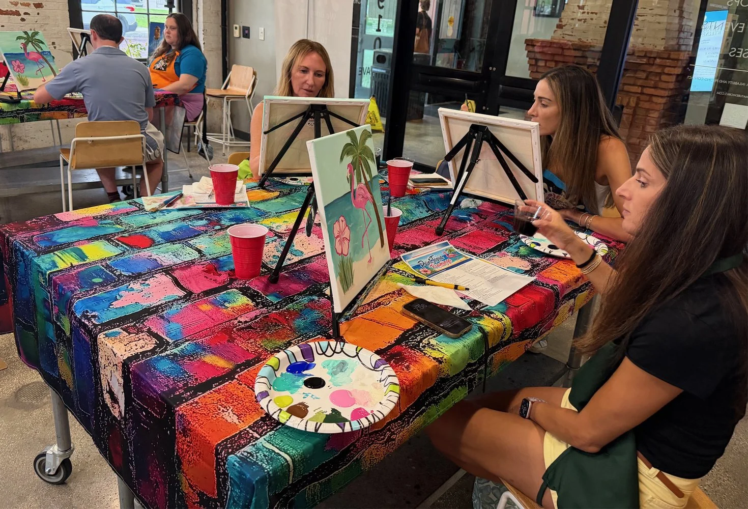 Women participating in a paint-and-sip art class at a decorated table with a colorful tablecloth, easels, canvases, and paint supplies, with a woman in the background looking at her phone.