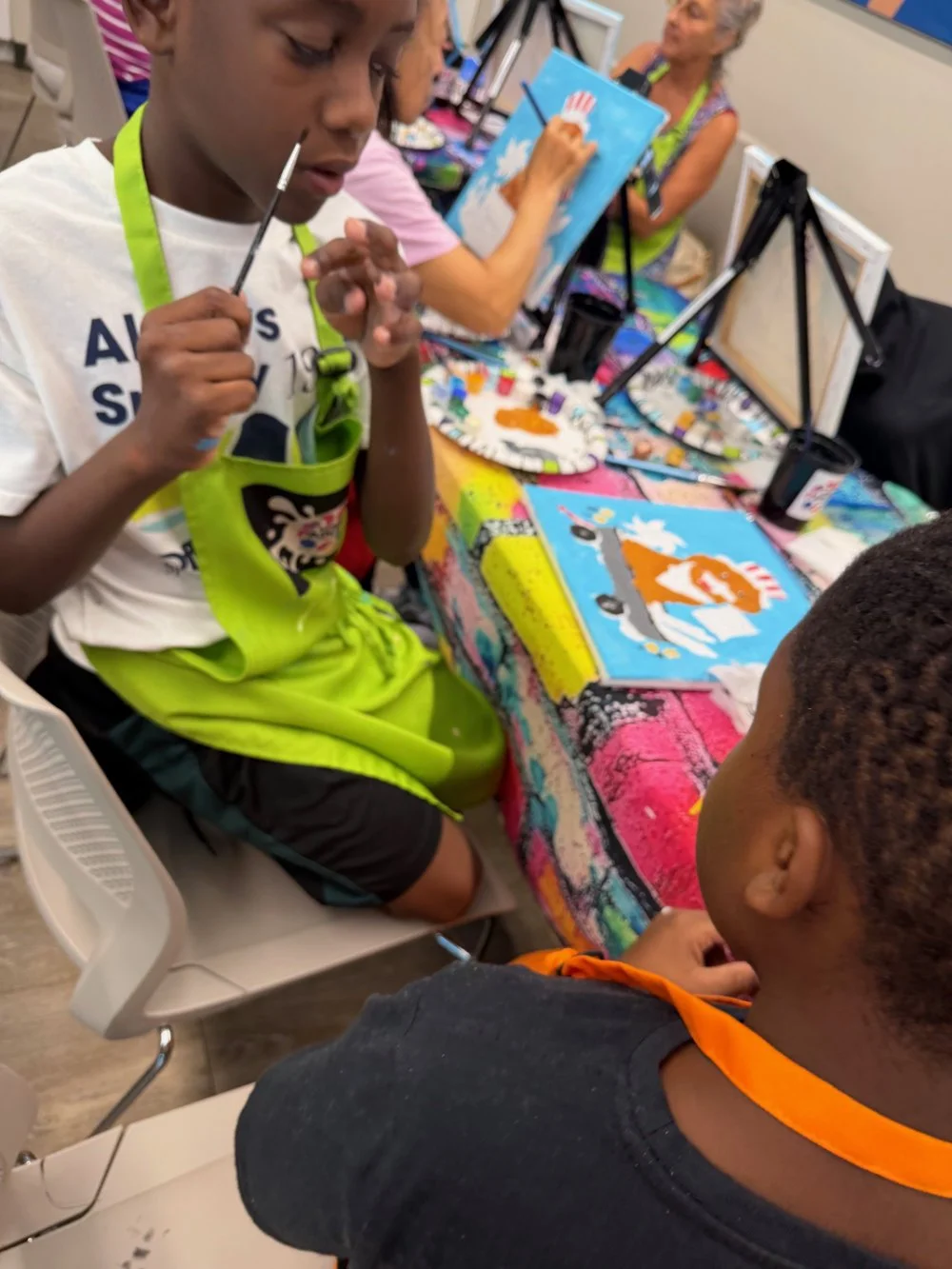 Children participating in a painting activity at a classroom or art workshop. They are creating pictures of a Thanksgiving turkey with a bright blue sky background.