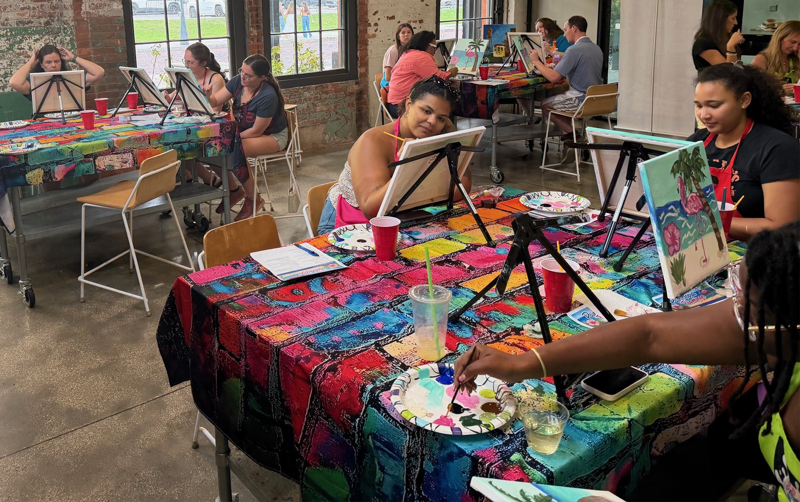 People participating in a painting class, seated at tables covered with colorful tablecloths, working on individual canvas paintings, with art supplies and drinks on the tables, in a studio with large windows and brick walls.