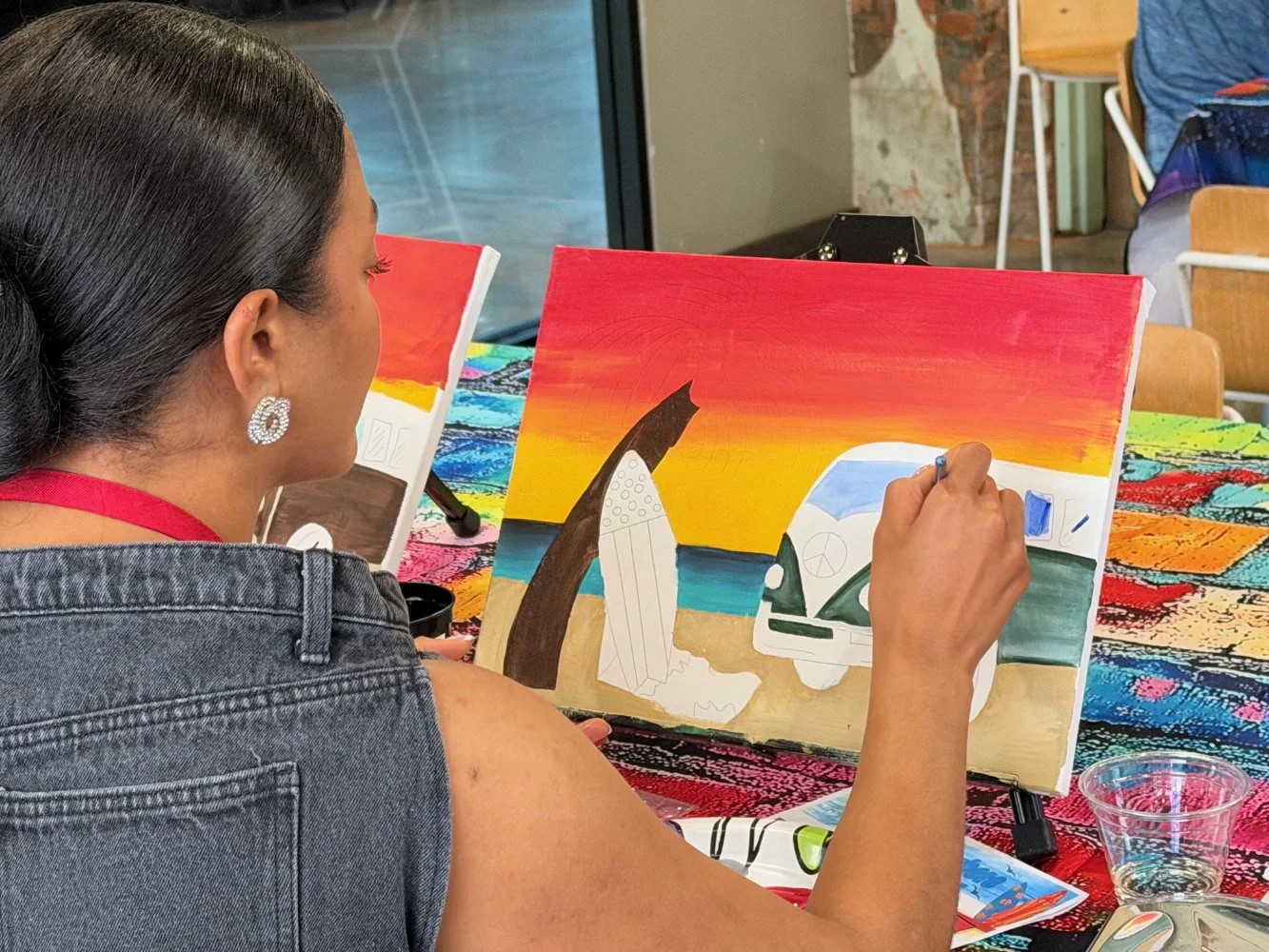 A woman with earrings and a denim jacket is painting a sunset scene on a canvas, depicting a colorful sky over the ocean with a white van and abstract shapes; she is seated at a table with art supplies and another canvas nearby.
