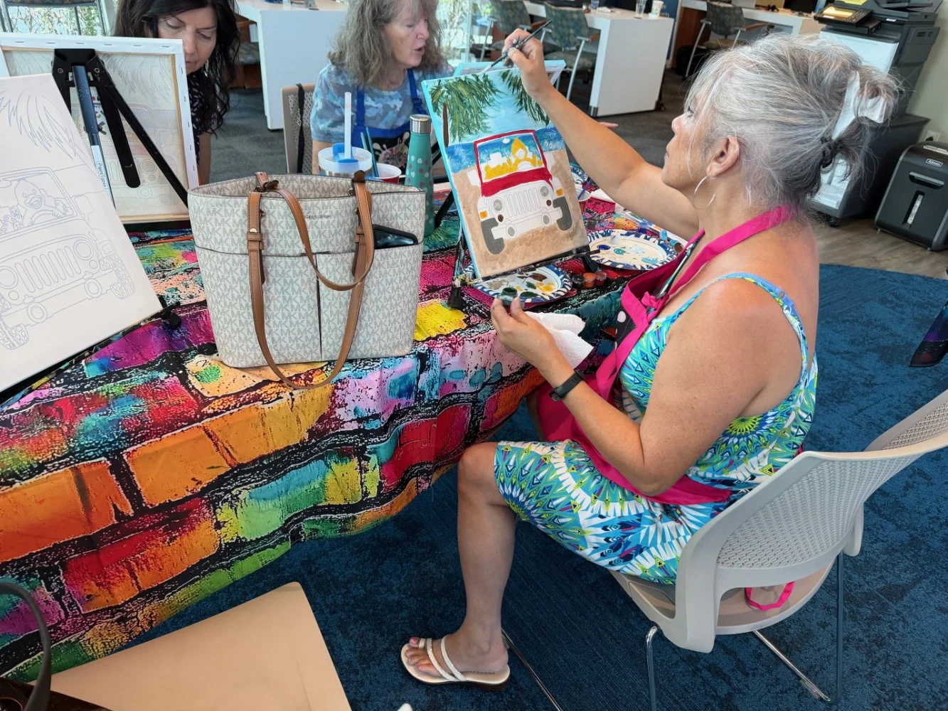 A woman with gray hair in a ponytail, wearing a colorful dress and pink apron, is painting a picture of a jeep on an easel at a colorful table. Other women are visible working on their paintings in the background.