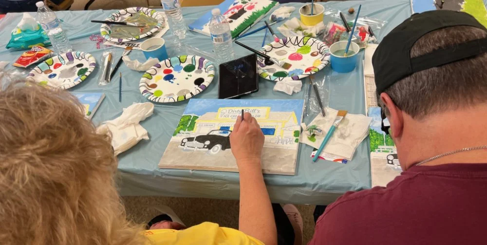 People painting a scene of a car dealership building on canvas at a table with discarded paper plates, water bottles, and painting supplies.