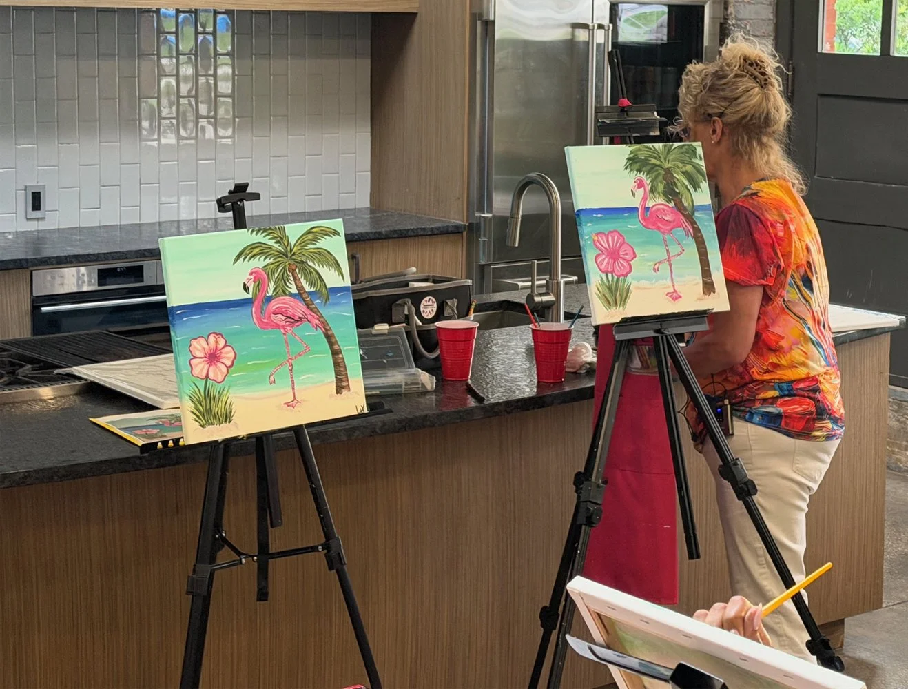 A woman painting a beach scene with a pink flamingo, palm tree, and hibiscus flower on a canvas in a kitchen-style studio.