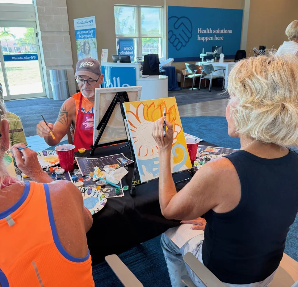 A group of middle-aged women and a man painting at a table in an indoor community center. They are creating colorful artwork with paint. The background features a banner with a handshake icon and the words "Health solutions happen here," and another 