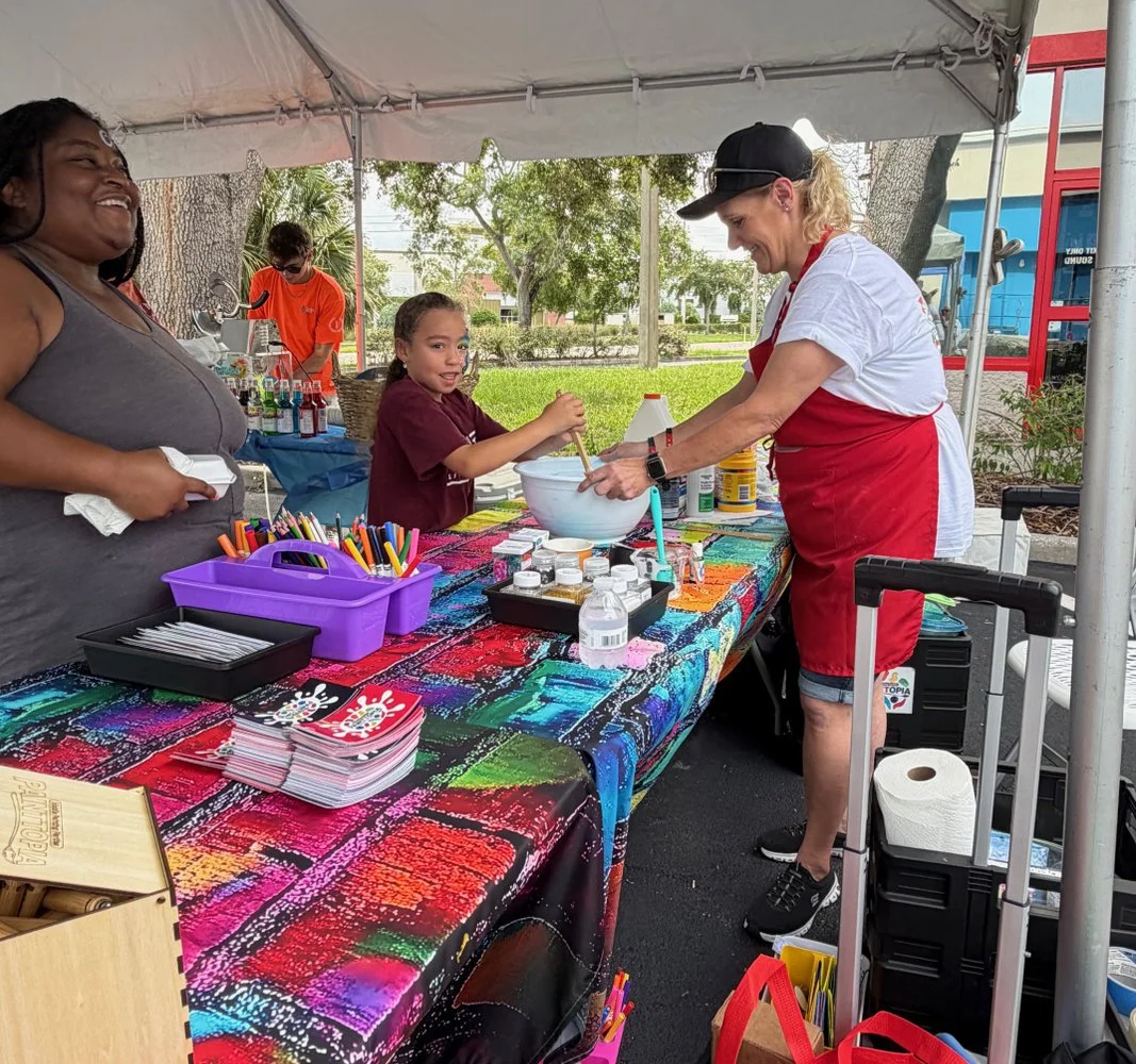 A woman with dark hair and a gray tank top laughs at a booth with colorful art supplies, while a young girl with a ponytail and a maroon shirt stirs a large white bowl. A woman in a white shirt, red apron, and black cap assists her. The booth is set 