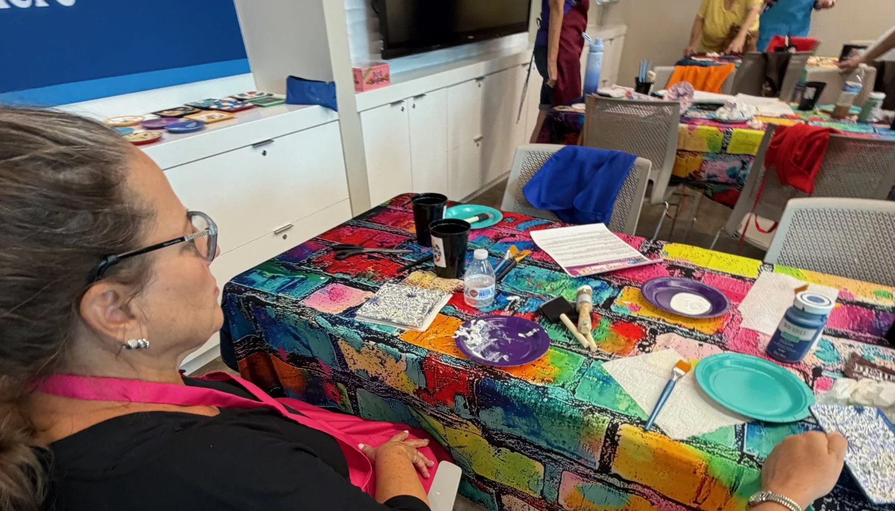 A woman with glasses and earrings sitting at a colorful table with art supplies, paper scraps, and cups, surrounded by other tables and people in a craft or art workshop setting.