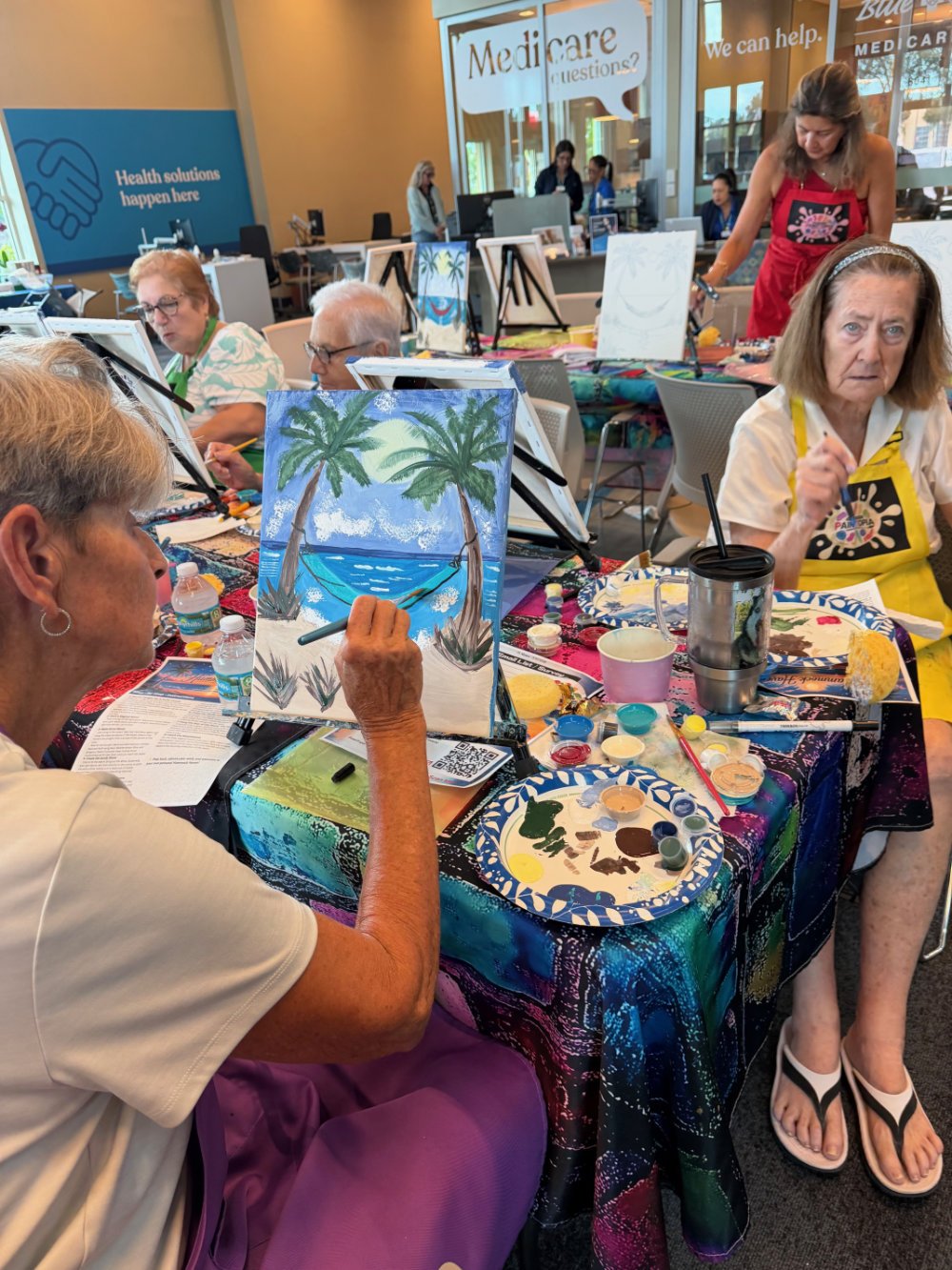 Senior women painting at an art class, with a tropical beach scene featuring palm trees and a hammock depicted on one canvas. The table is covered with art supplies, water bottles, and cups.