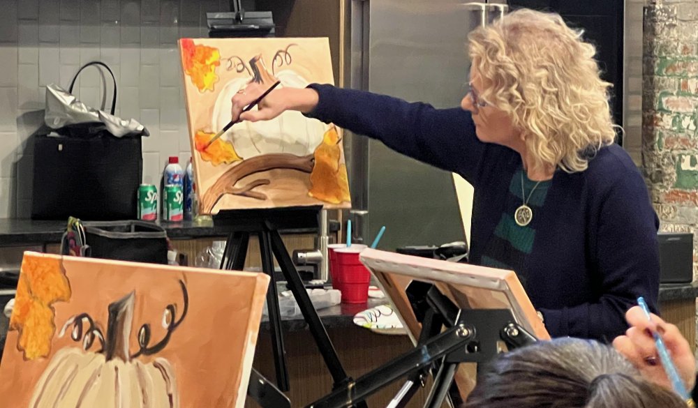 Woman with curly blonde hair painting a fall-themed picture of a pumpkin with leaves on an easel.