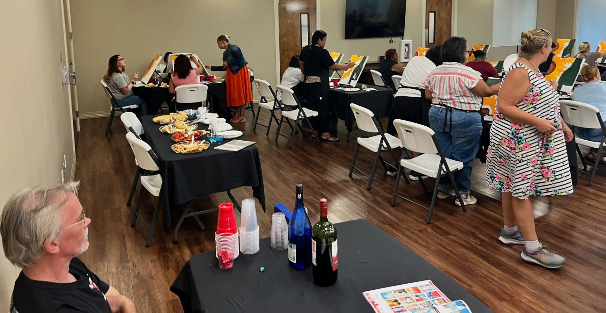 People gathered at a social event with food, drinks, and painting activities in a room with wooden floors and beige walls.