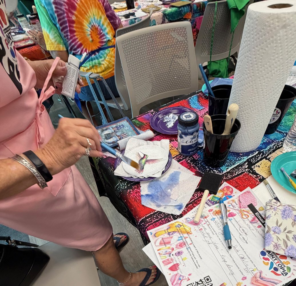 A person is painting a small object, possibly a figurine, on a table covered with colorful and patterned tablecloths. The table has various art supplies like brushes, paper towels, and markers. Other tables with similar setups are visible in the back