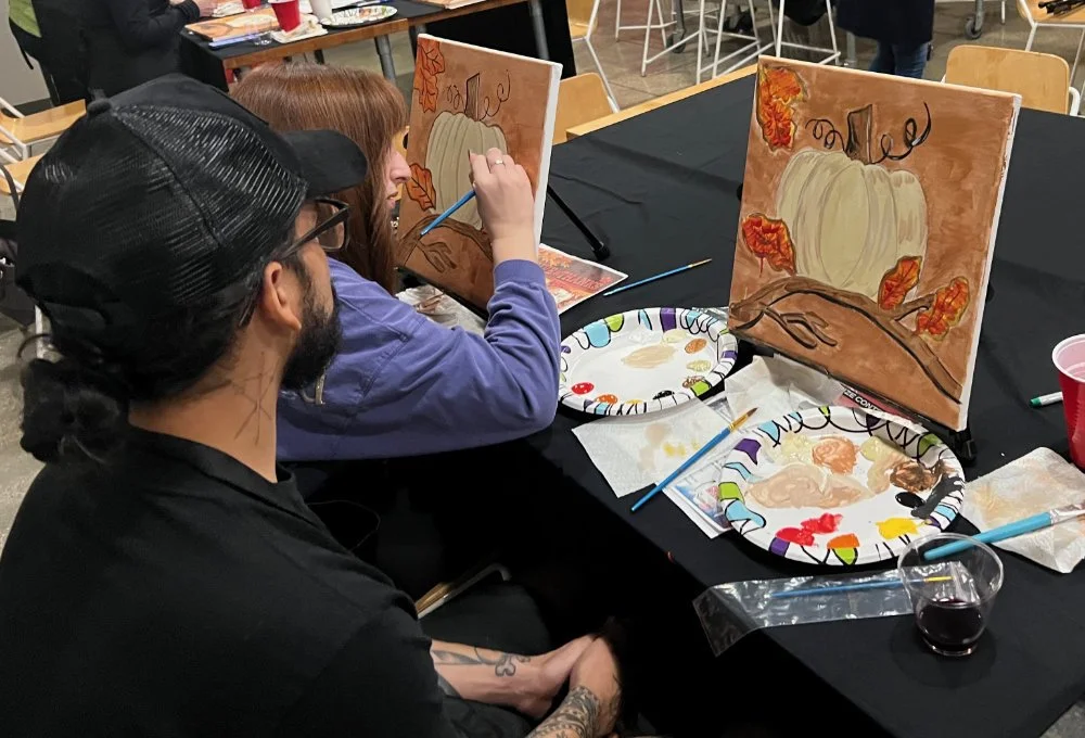 People painting pumpkins on canvases at a table during a painting event.