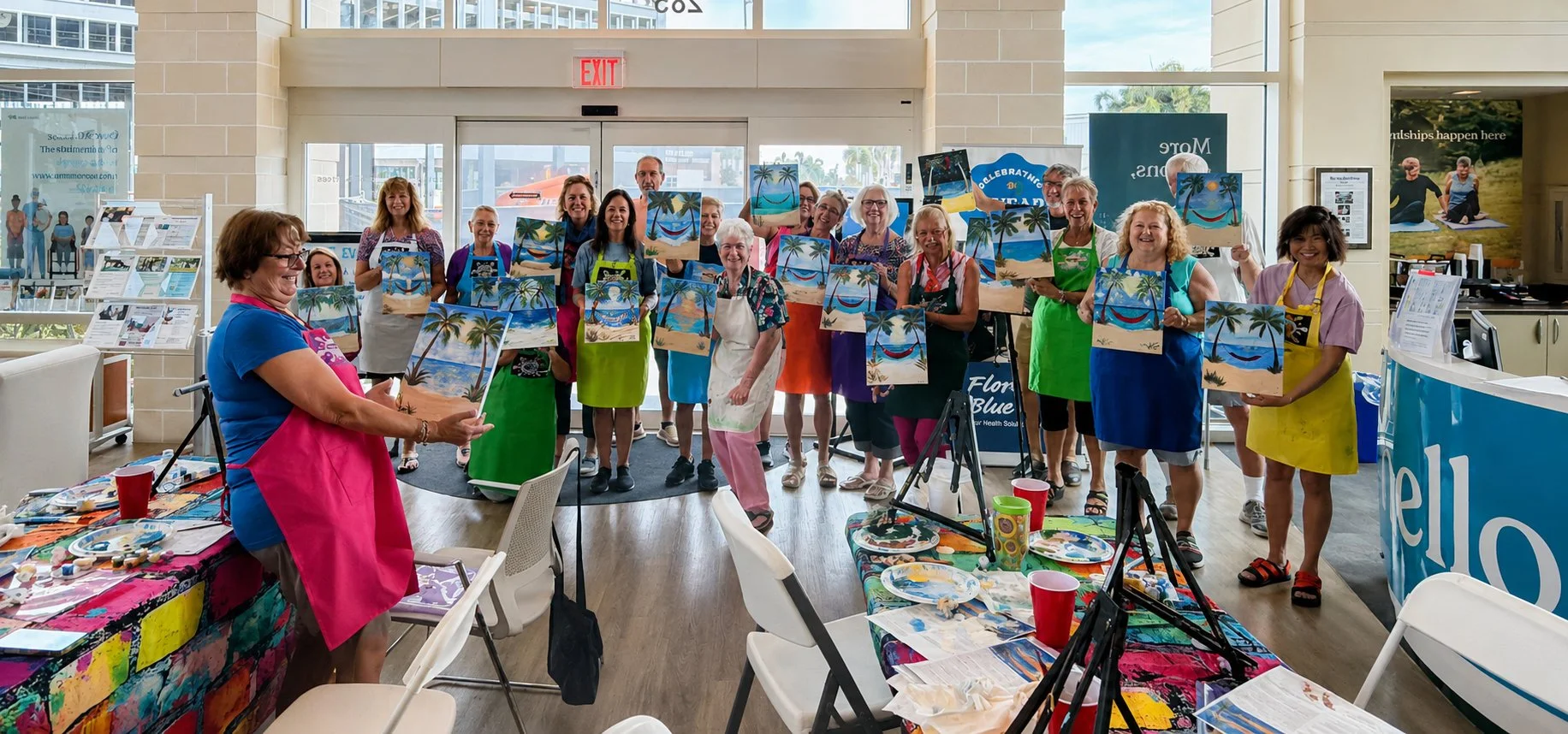 Group of people, mostly women, holding up painted canvases with beach and palm tree scenes at an art class event in a well-lit indoor space, with art supplies and paintings visible.