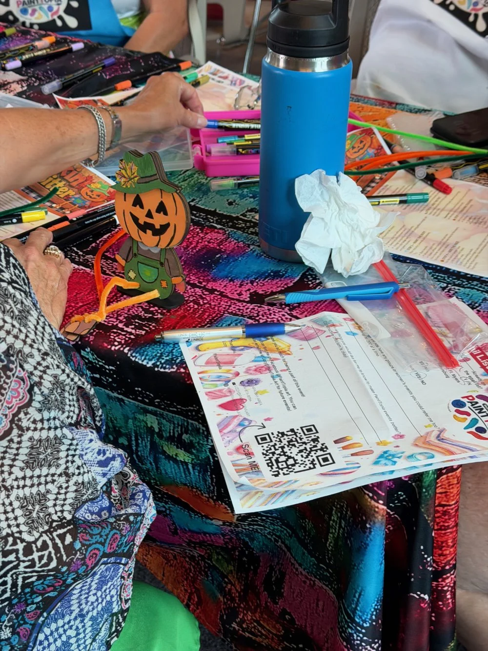 Decorative Halloween-themed craft table with colorful markers, a blue water bottle, a tissue, a paper with a QR code, and a Halloween jack-o'-lantern figure at the center.
