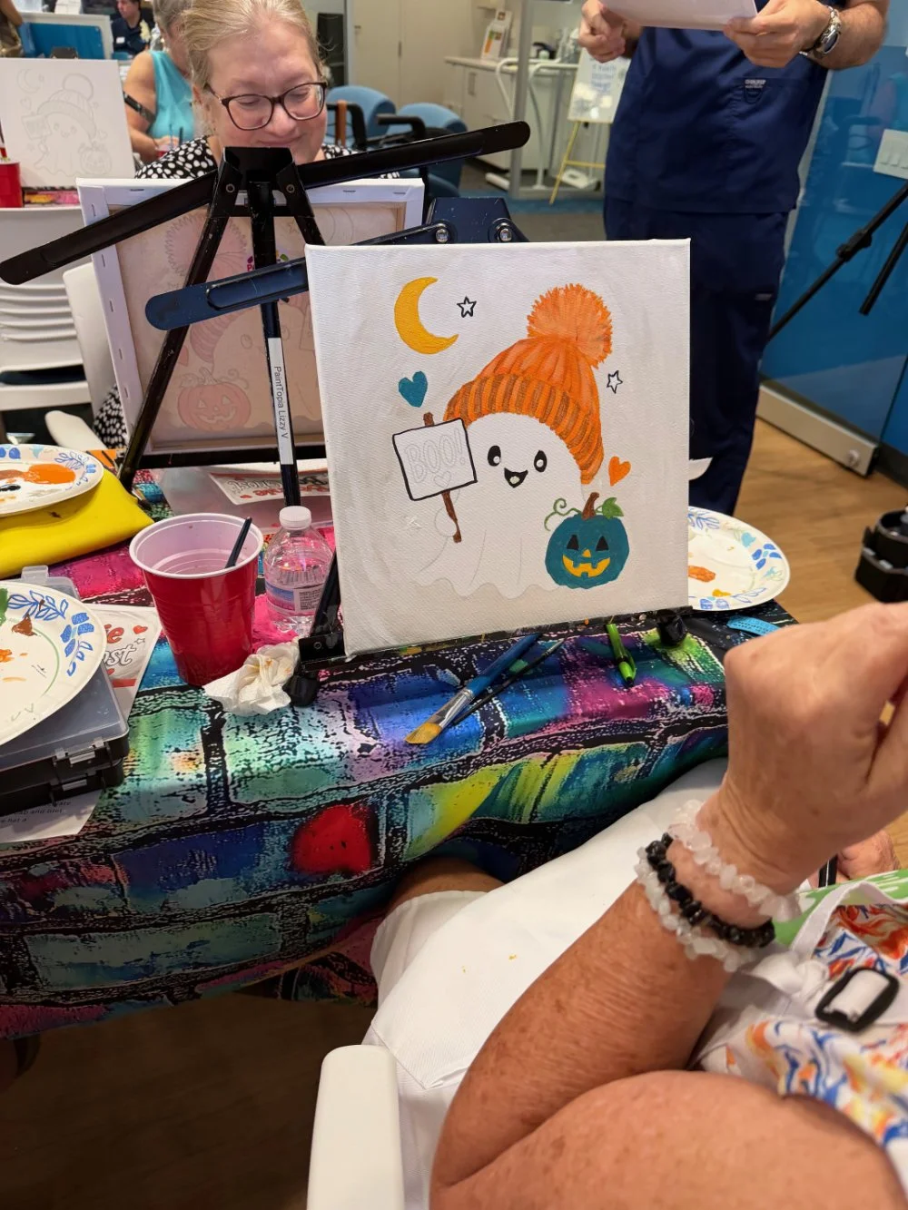 A person is painting a Halloween-themed illustration featuring a smiling ghost wearing an orange knit hat, holding a sign that says 'BOO!', next to a jack-o'-lantern pumpkin, with a purple and orange background and small colorful hearts and stars. Th