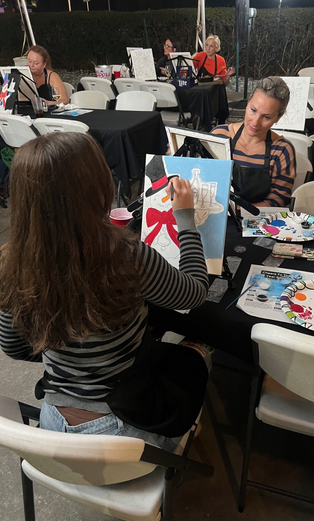 Women participating in a paint and sip event, painting holiday-themed artwork featuring a wine bottle, glasses, and a red bow, seated at tables with painting supplies and drinks, outdoor setting at night.