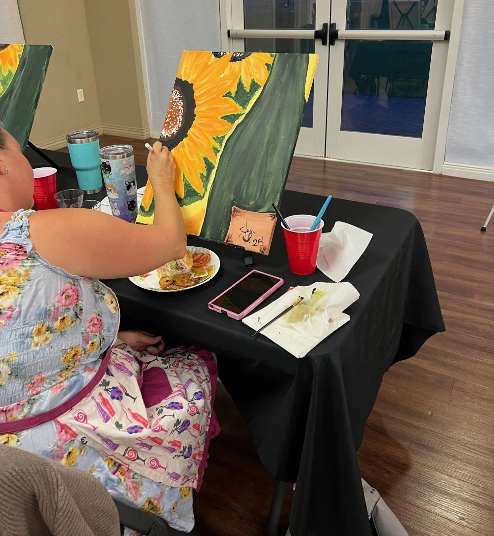 Person painting a sunflower on a canvas at a table with art supplies and food, in an indoor setting.