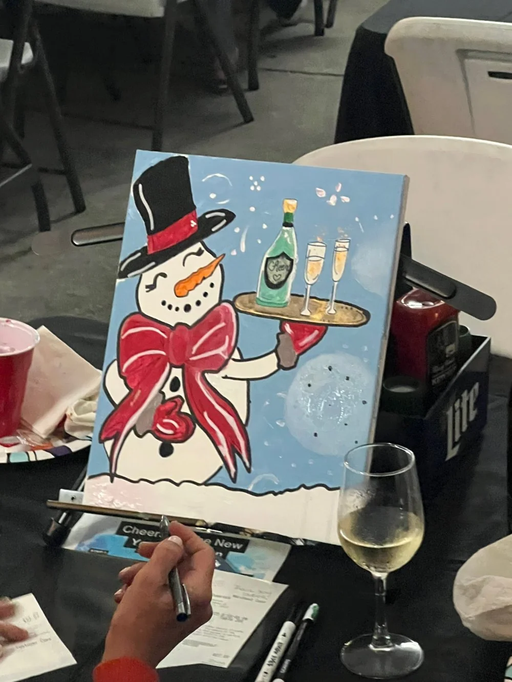 A person is painting a snowman on a canvas with a bottle of champagne, two glasses of champagne, and a wine glass of white wine on the table. The painting depicts a snowman with a top hat, orange nose, and red scarf, holding a tray with champagne and
