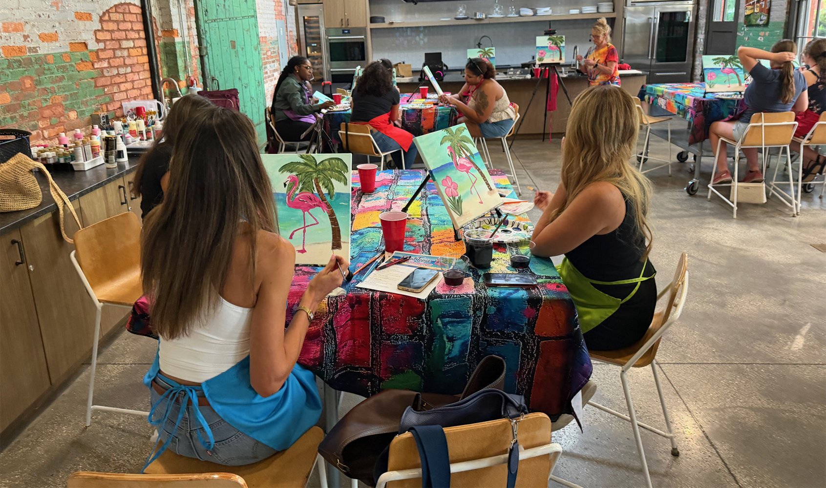 A group of women participating in a paint and sip class inside a creative studio with a brick wall and a kitchen in the background. They are painting tropical-themed pictures featuring pink flamingos and palm trees on canvases set on a colorful table
