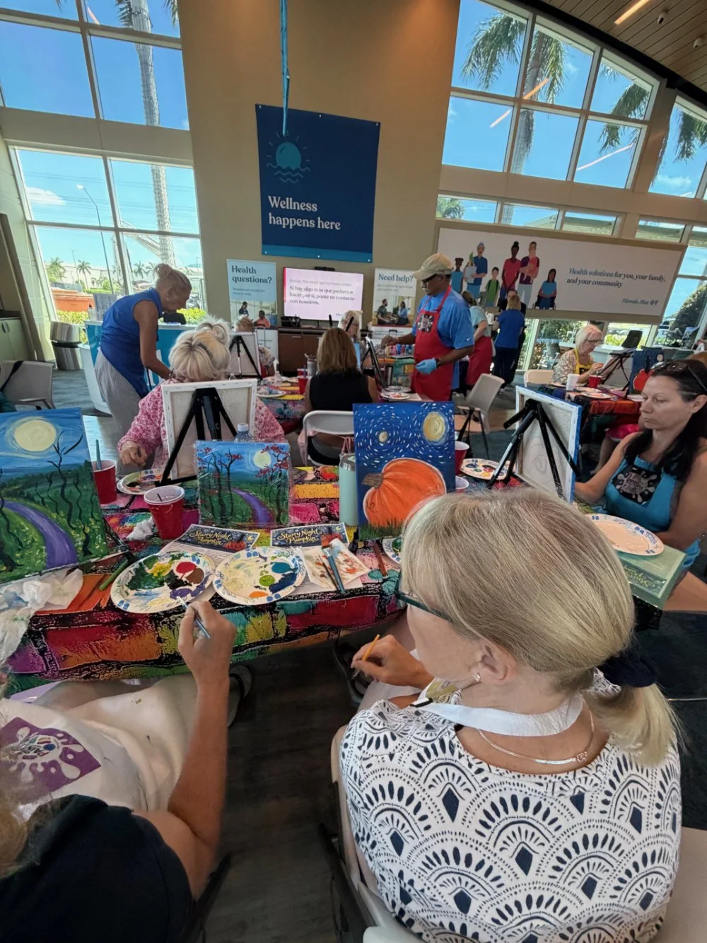 People participating in a group painting activity at a community event inside a large room with tall windows, palm trees, and a blue sky outside. Art supplies and finished paintings, mostly of outdoor scenes and pumpkins, are on the tables.