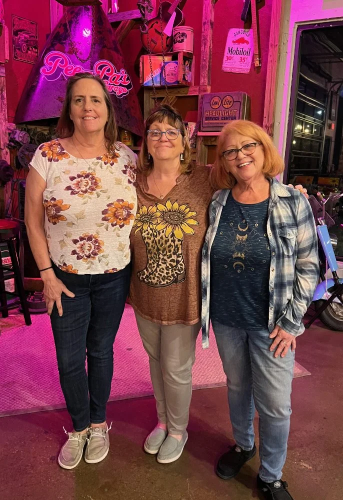 Three women standing inside a vintage garage or workshop with pink lighting, surrounded by retro signs and motorcycle parts.