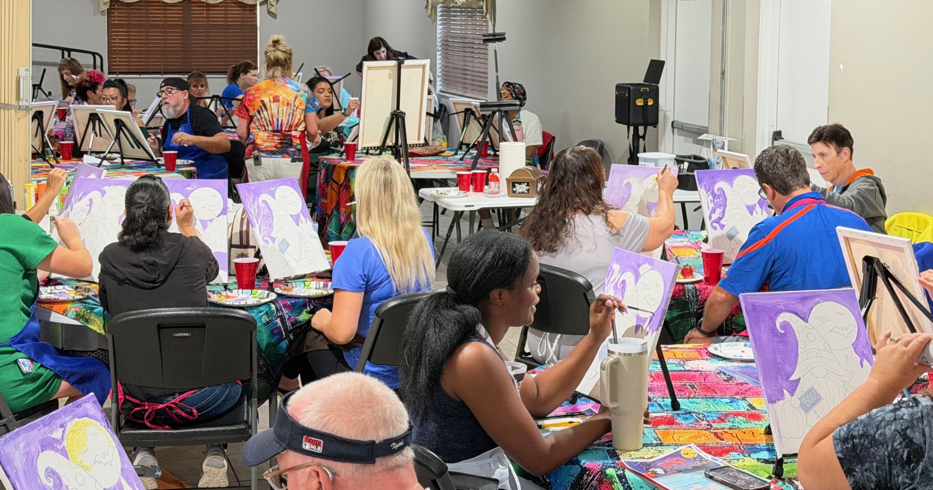 People participating in a group painting class, creating pictures of purple and white swan in a room with tables, easels, and art supplies.