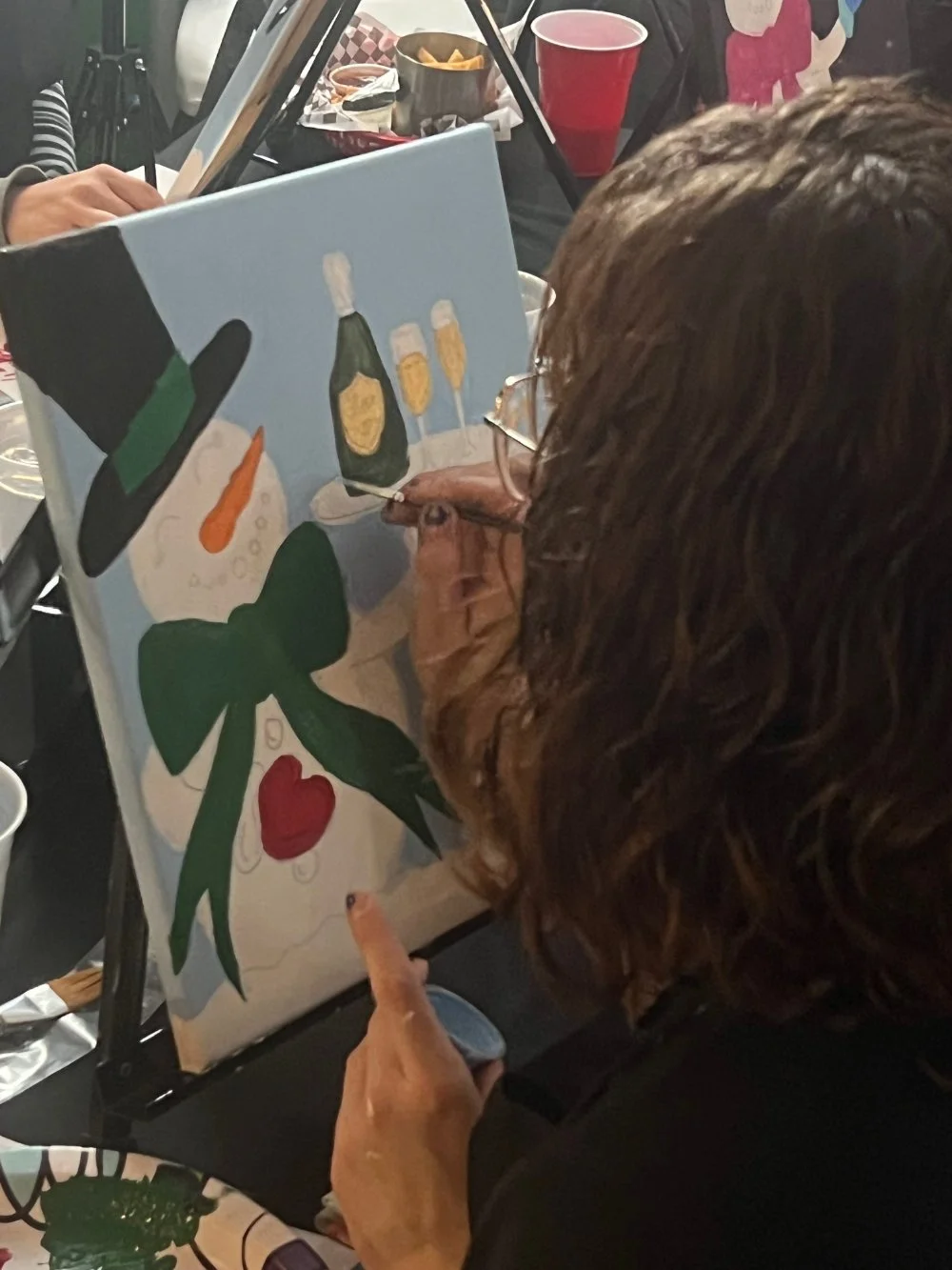 Person painting a holiday-themed canvas with a snowman wearing a black top hat, an orange carrot nose, a green scarf, and a red heart on the snowman's body. There are also bottles of champagne and glasses in the background, with trays of snacks and c