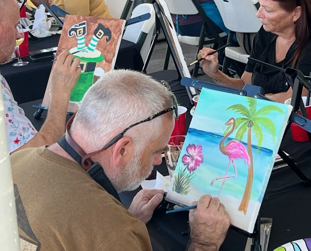 People painting colorful pictures, one of a flamingo standing near palm trees and a hibiscus flower.