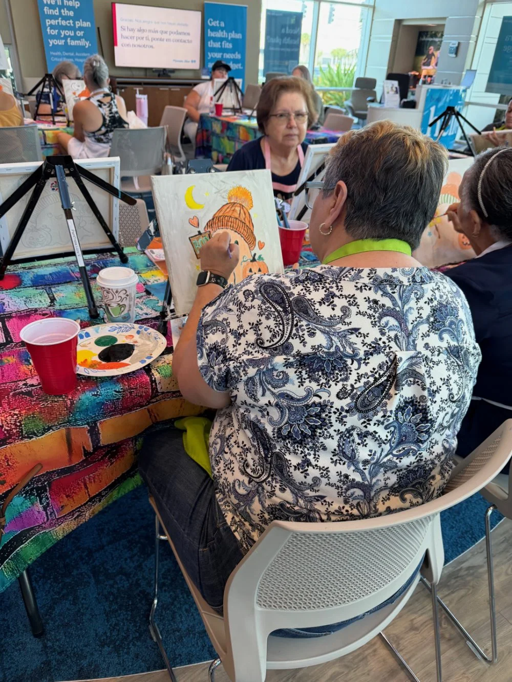 A group of people participating in a painting activity at a colorful table, with each person working on their own artwork. In the foreground, a woman with short hair and glasses is painting a fall-themed picture with a pumpkin and trees, while others