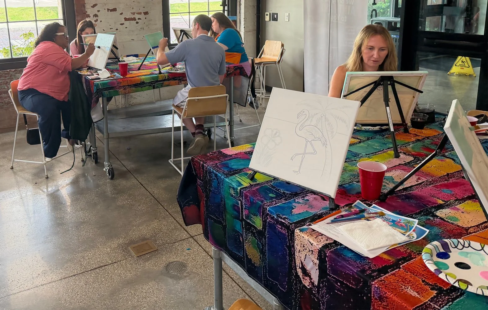People participating in a painting class, sitting at tables covered with colorful tablecloths, painting on canvases, with a large window in the background.