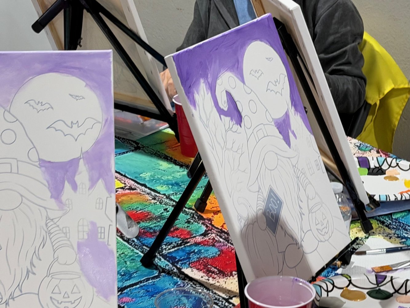 Halloween-themed painting setup with canvases, a rainbow tablecloth, and paints. One canvas features a halloween scene with a pumpkin, haunted house, moon, and bats, outlined in pencil and partially painted purple.