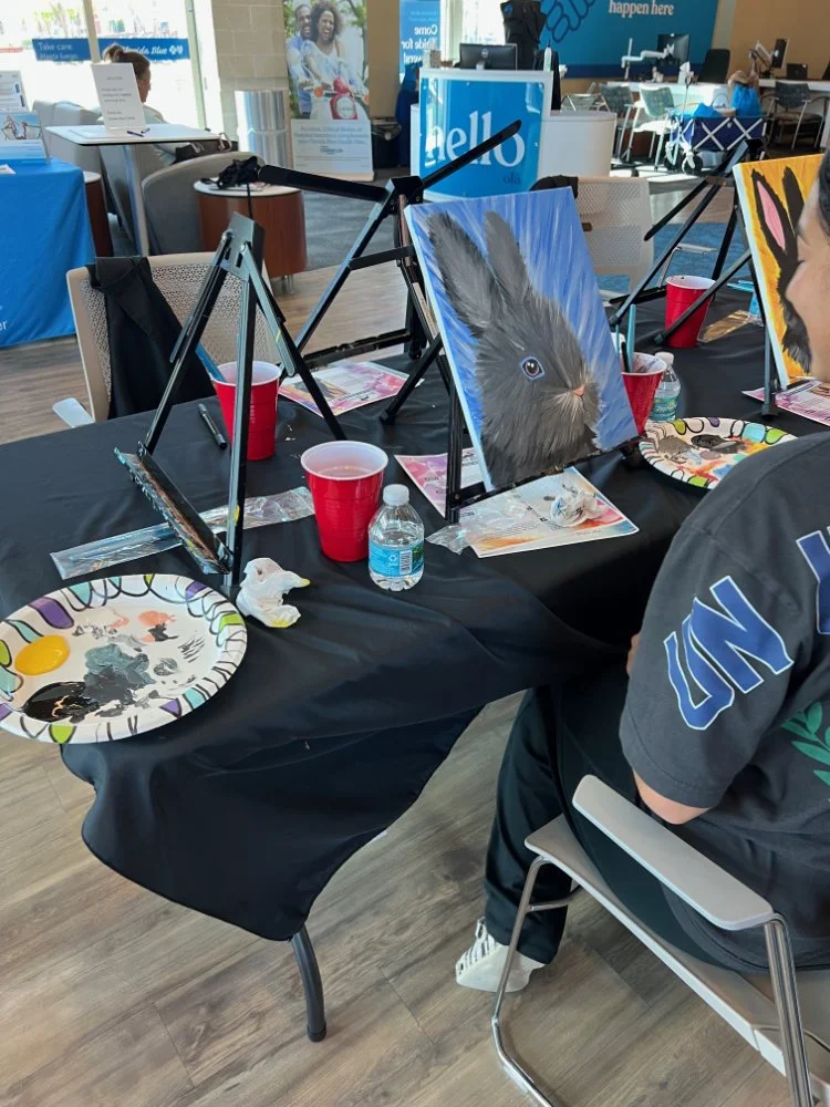 Paintings of animals, including a detailed bunny and a dog, are displayed on easels at a table during an art event. Art supplies, cups, and water bottles are also on the table.