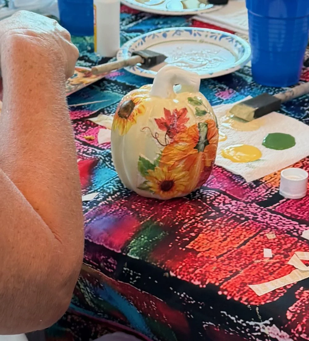A pumpkin painted with fall-themed designs, surrounded by painting supplies on a colorful tablecloth.