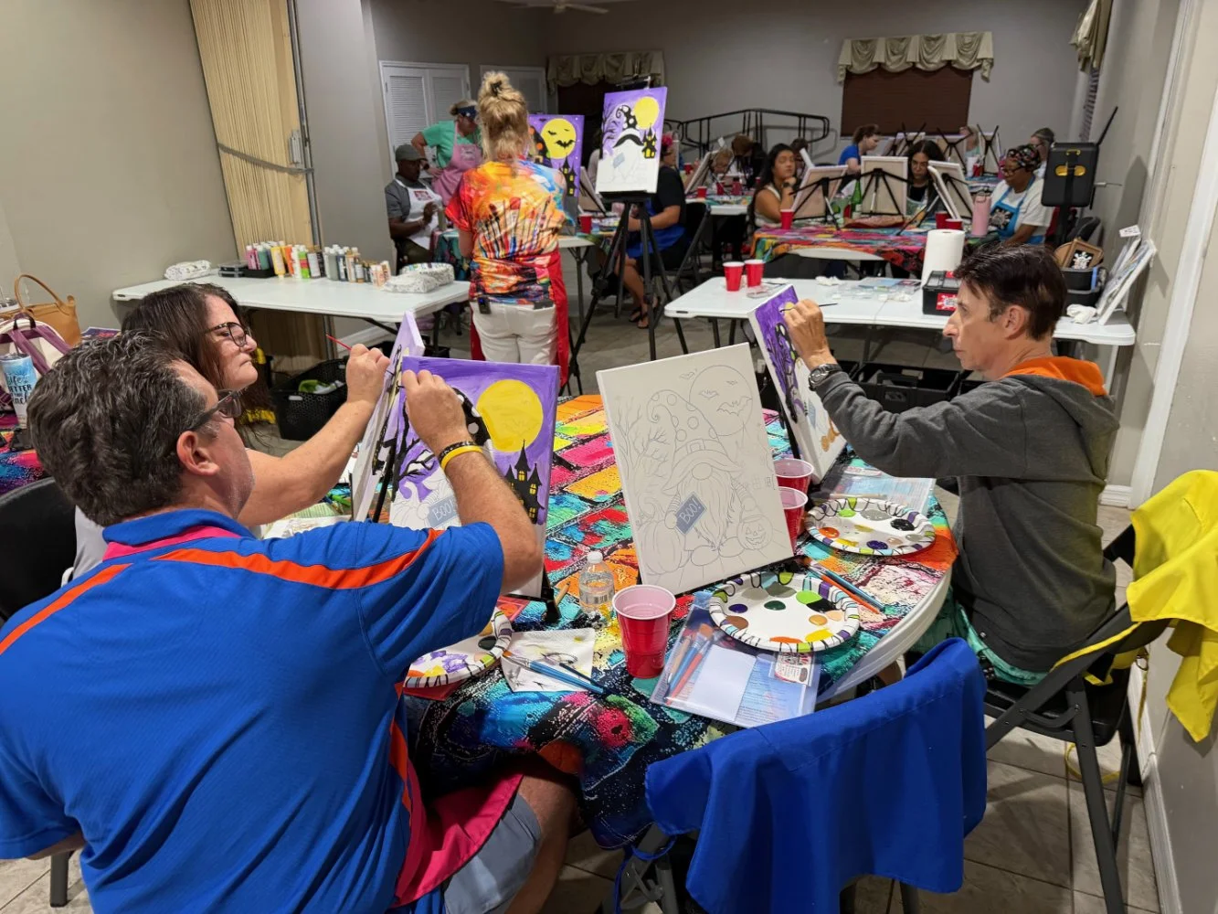 People painting Halloween-themed pictures at a group art event in a room decorated for Halloween.