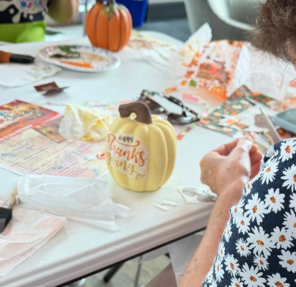 A table decorated for Thanksgiving with a ceramic pumpkin candle that says 'Happy Thanksgiving,' paper napkins, and scattered paper decorations, with a person sitting nearby wearing a daisy-patterned shirt.