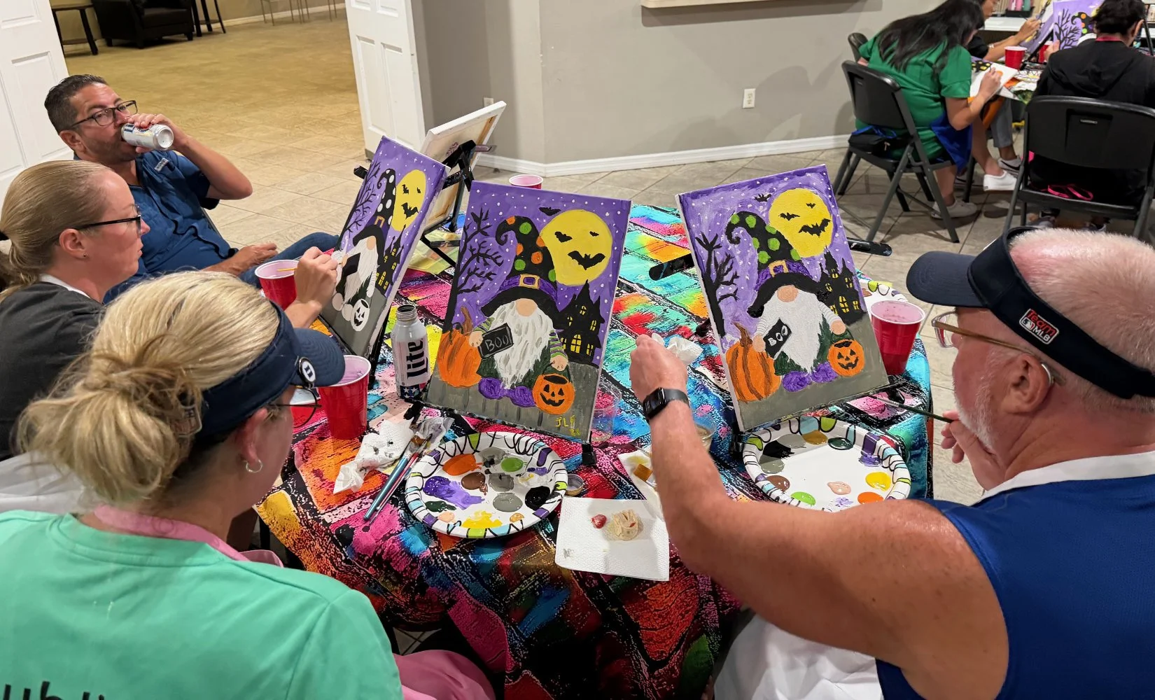 Group of people participating in a Halloween-themed painting activity. They are painting pumpkins, witches, and spooky scenes on canvases at a table covered with colorful paint splashes. The scene includes painting supplies, drinks, and several compl