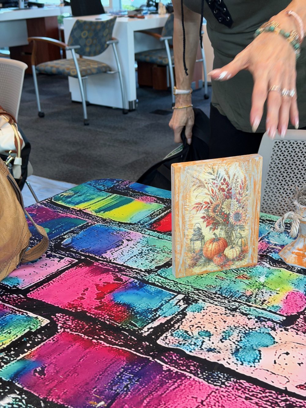 Tables covered with colorful, abstract-patterned cloths, with a small painting of a floral arrangement and pumpkins on one table, inside a room with chairs and a counter in the background.