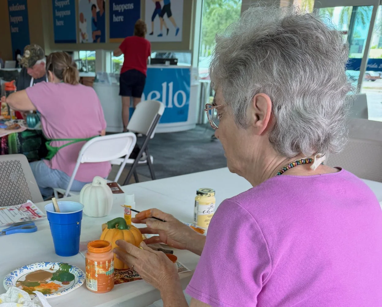 Older woman painting a small pumpkin at a craft table in a community center, with other people engaged in activities in the background.
