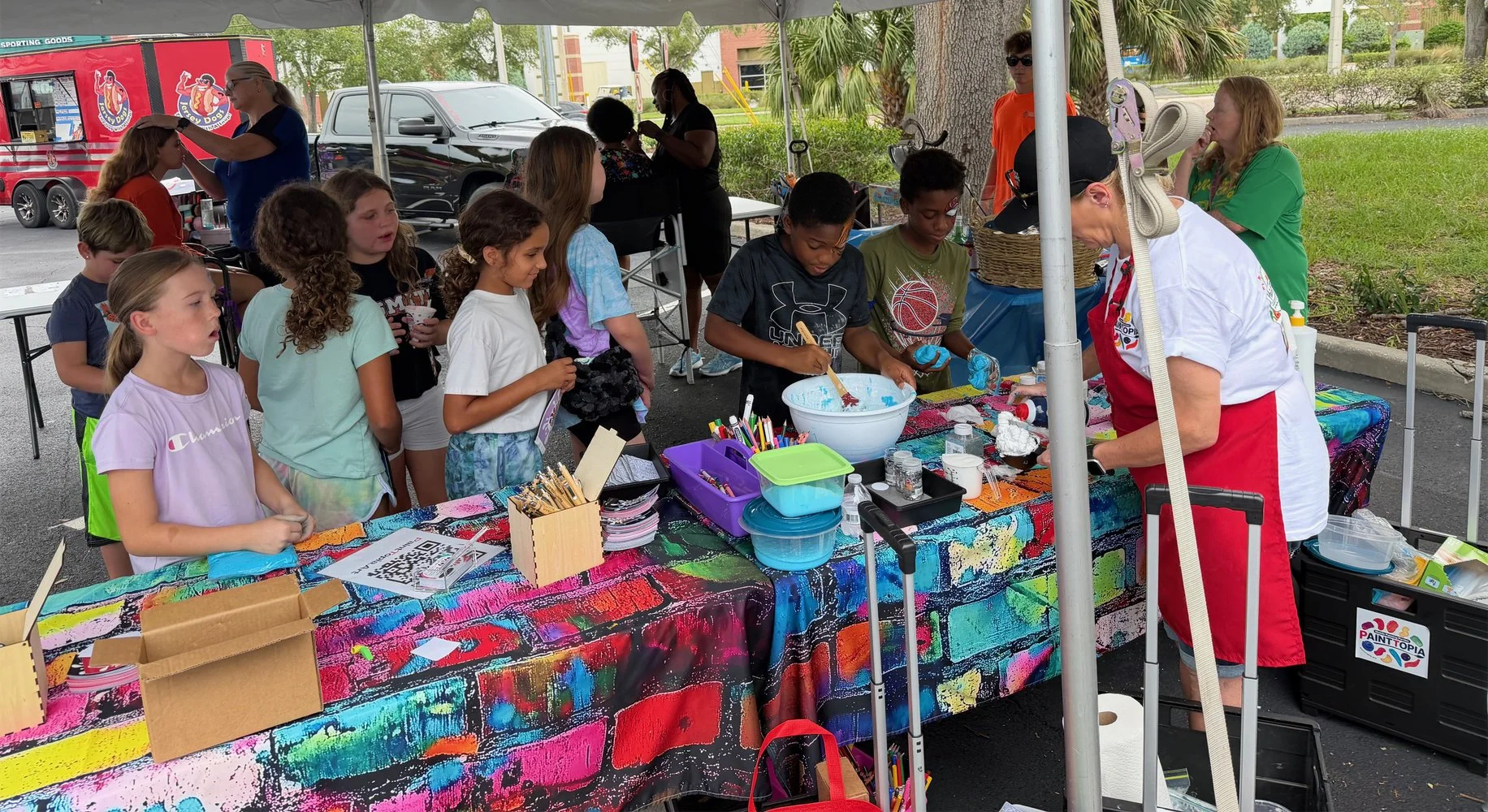 Children gathered around a table at a craft booth, engaging in a painting activity with colorful supplies, under a white canopy outdoors. Adults are supervising, with some preparing or assisting at the table.