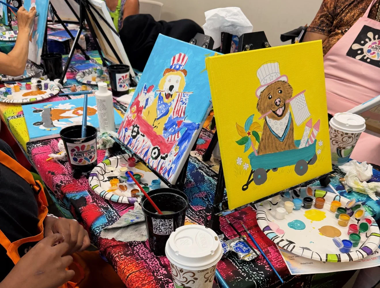 Room with people painting animal-themed artworks on canvas, with paints, brushes, and cups on a colorful table.