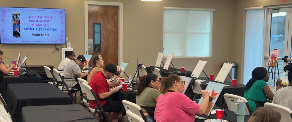 People participating in a paint and sip class, sitting at tables with canvases and paint supplies, while a large screen displays event details.