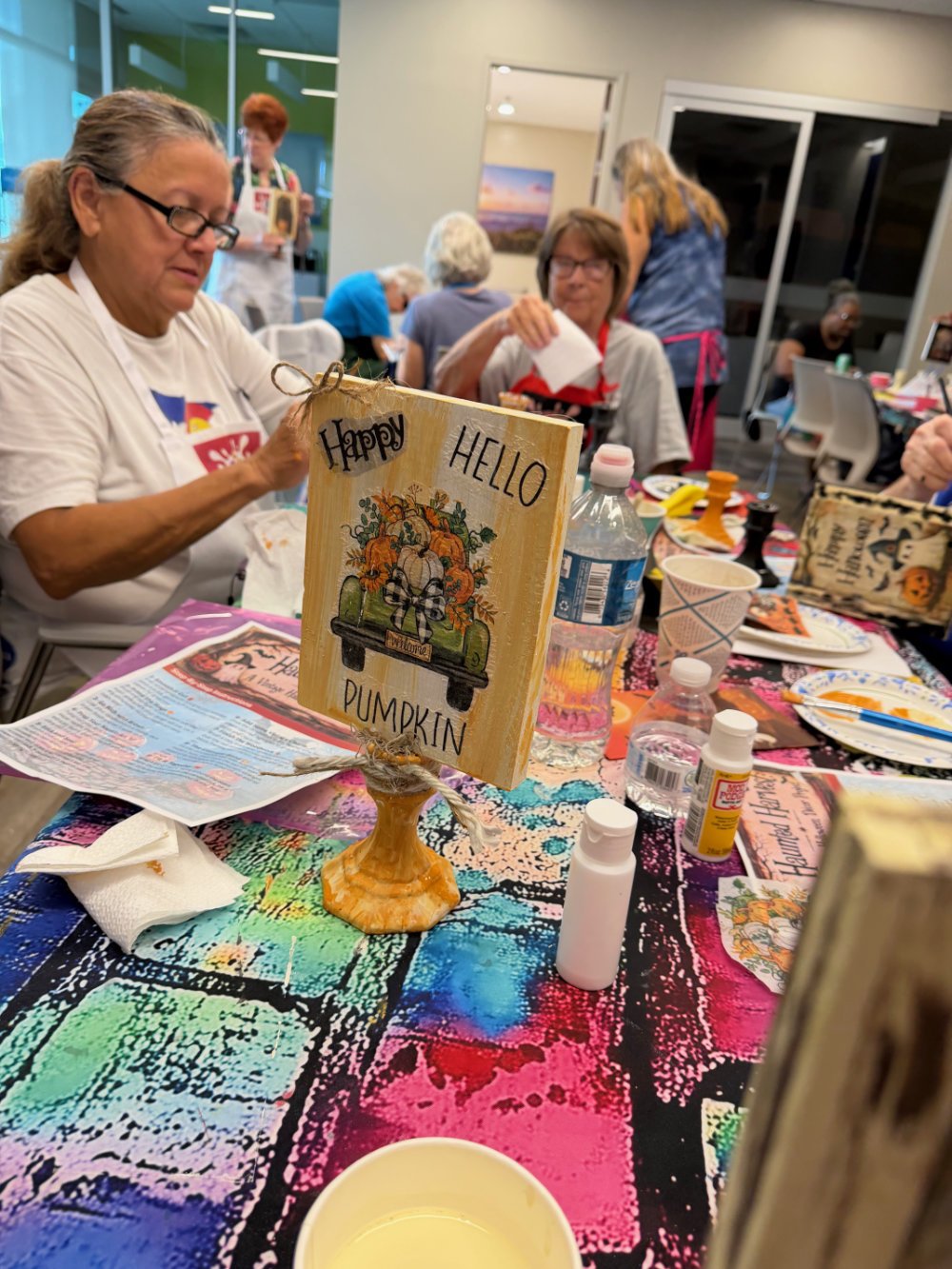 Group of women sitting around a table decorated for a gathering, with a sign that says 'Happy Hello Pumpkin' and various craft supplies and drinks on the table.
