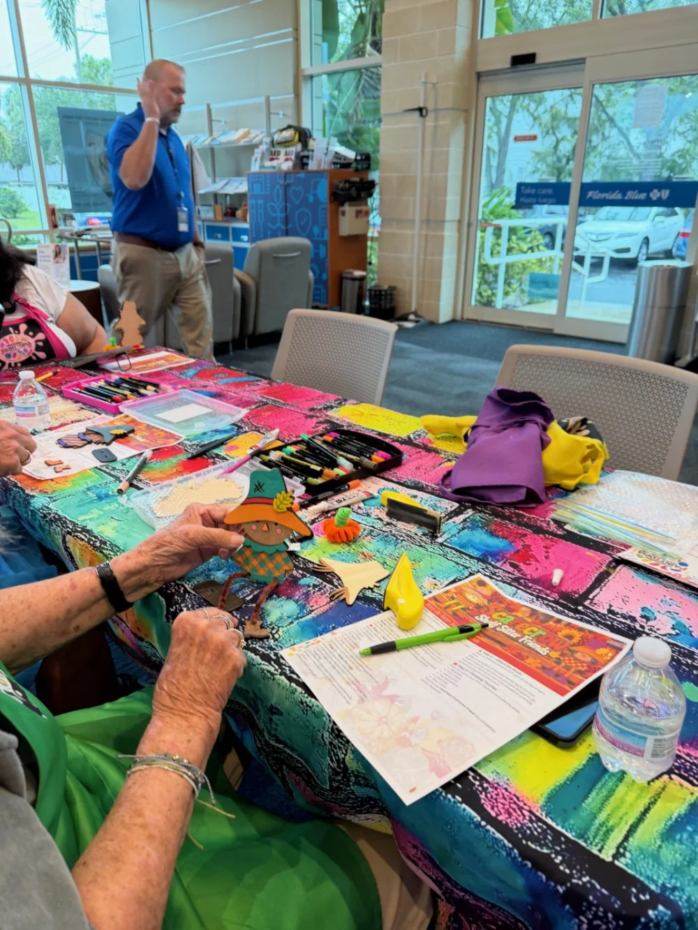 People participating in a craft activity at a table with colorful decorations and supplies, inside a building with large windows.