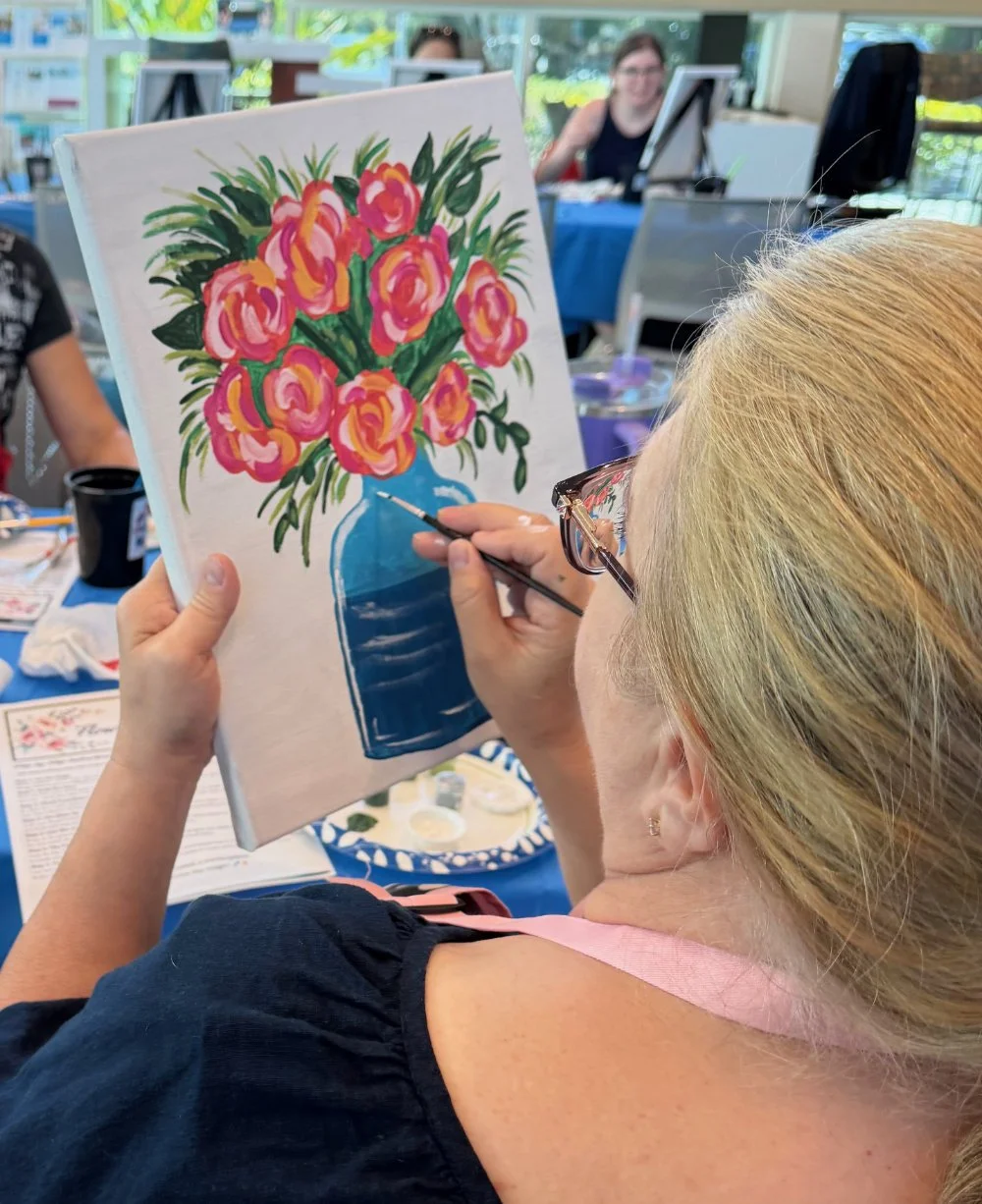Woman painting a colorful bouquet of pink and orange roses in a blue vase on a canvas at a painting class.