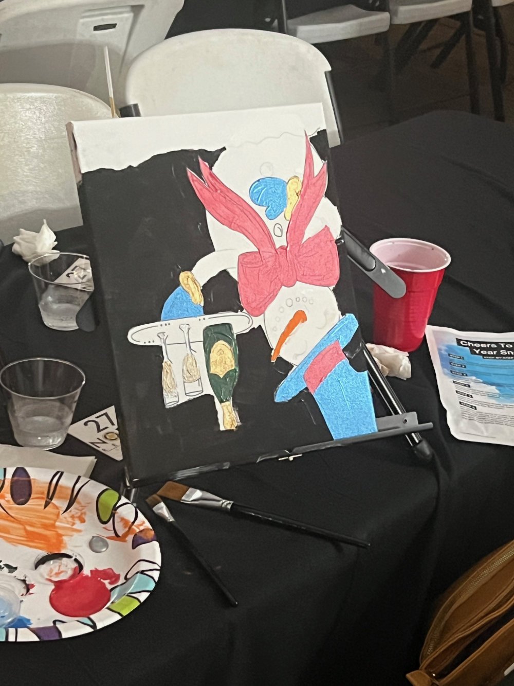 A painting of a snowman with a blue nose, red scarf, and red wings, sitting at a table with a bottle of champagne, alongside a wine glass, and surrounded by party supplies on a black table.