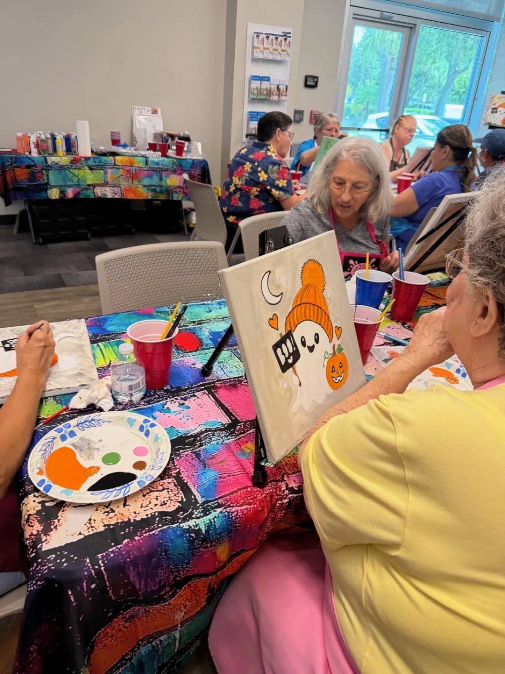Group of elderly women participating in a Halloween-themed painting activity in a room with large windows. They are seated at a table covered with a colorful tablecloth, working on ghost and pumpkin paintings. Art supplies and drinks are on the table