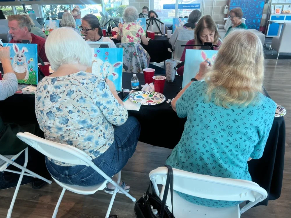 Group of people participating in a painting class, painting Easter-themed bunny pictures on canvases in a bright room.