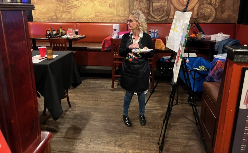 An elderly woman with curly gray hair, glasses, wearing a black apron and jeans, standing in a restaurant or cafe, holding a palette and brush, painting on a canvas set on an easel.