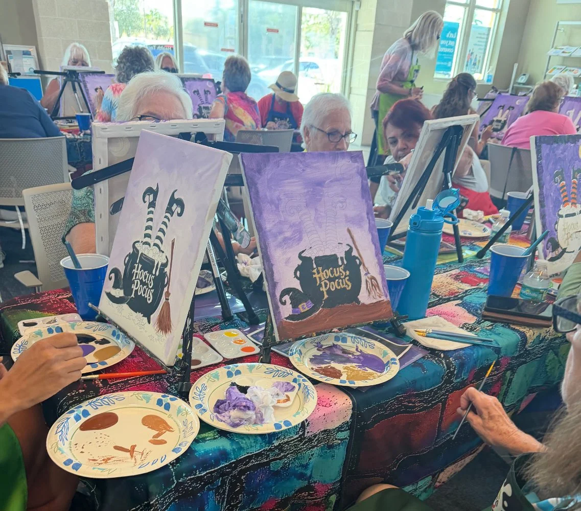 A group of people participating in a painting class themed 'Hocus Pocus' at a crowded indoor venue with large windows, some painting on canvases, and supplies such as paint palettes, brushes, and water bottles on the table.