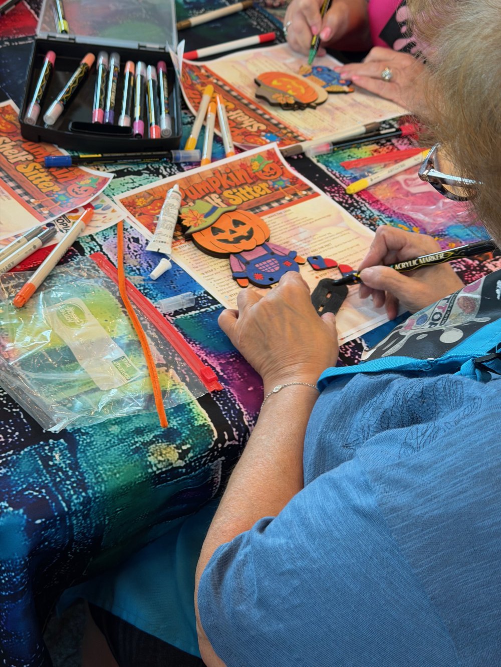 Person creating Halloween-themed paper crafts with markers, stickers, and cutouts on a colorful table.