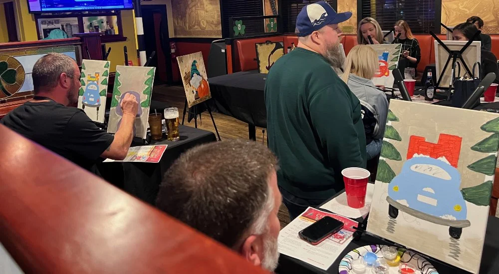People participating in a group painting activity at a restaurant, with painted canvases depicting holiday-themed scenes.